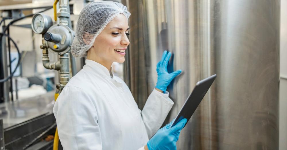 A worker in a white coat, hair net, and blue gloves, is smiling and reading a clipboard while touching a large metal tank.