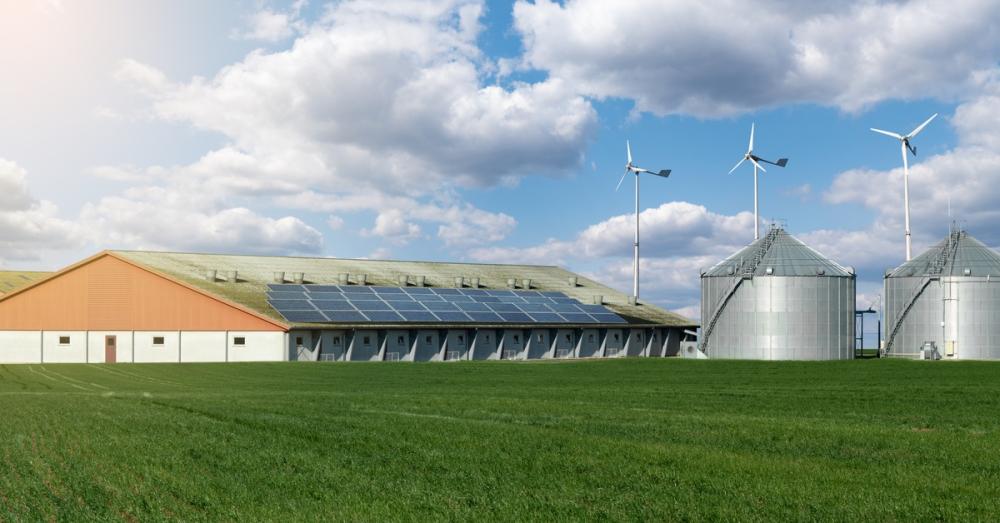 A farming facility with a large field of green crops, two grain silos, wind turbines, and solar panels on the roof.
