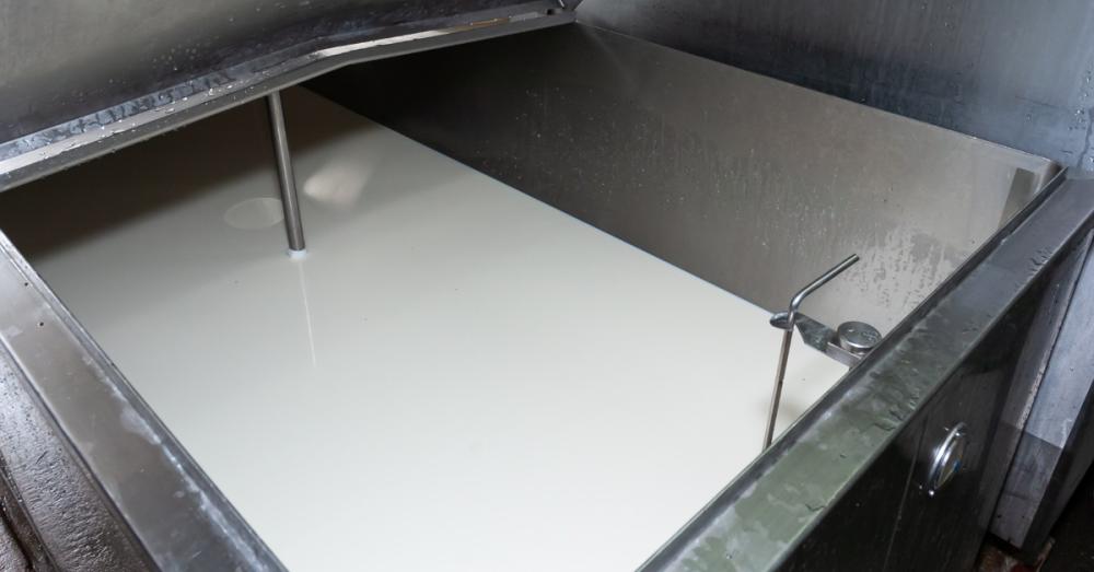 An open stainless-steel bulk milk tank is partially filled with milk. Metal supports and a hinged lid are visible.
