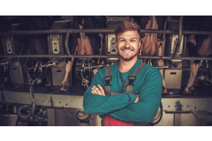 A man in a work apron and long-sleeved shirt smiling. He is standing in front of a row of cows in a miking parlor.