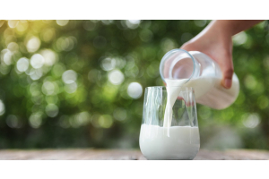 A hand pours milk from a glass bottle into a glass on a wooden table outdoors. Blurred greenery is in the background.