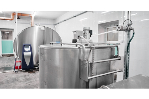 Two large stainless-steel milk tanks, pipes, hoses, motors, and control equipment sit inside a tiled processing room.