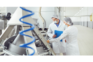 Two workers in white lab coats, masks, and hair nets are inspecting equipment. There is a blue, curled tube in the foreground.