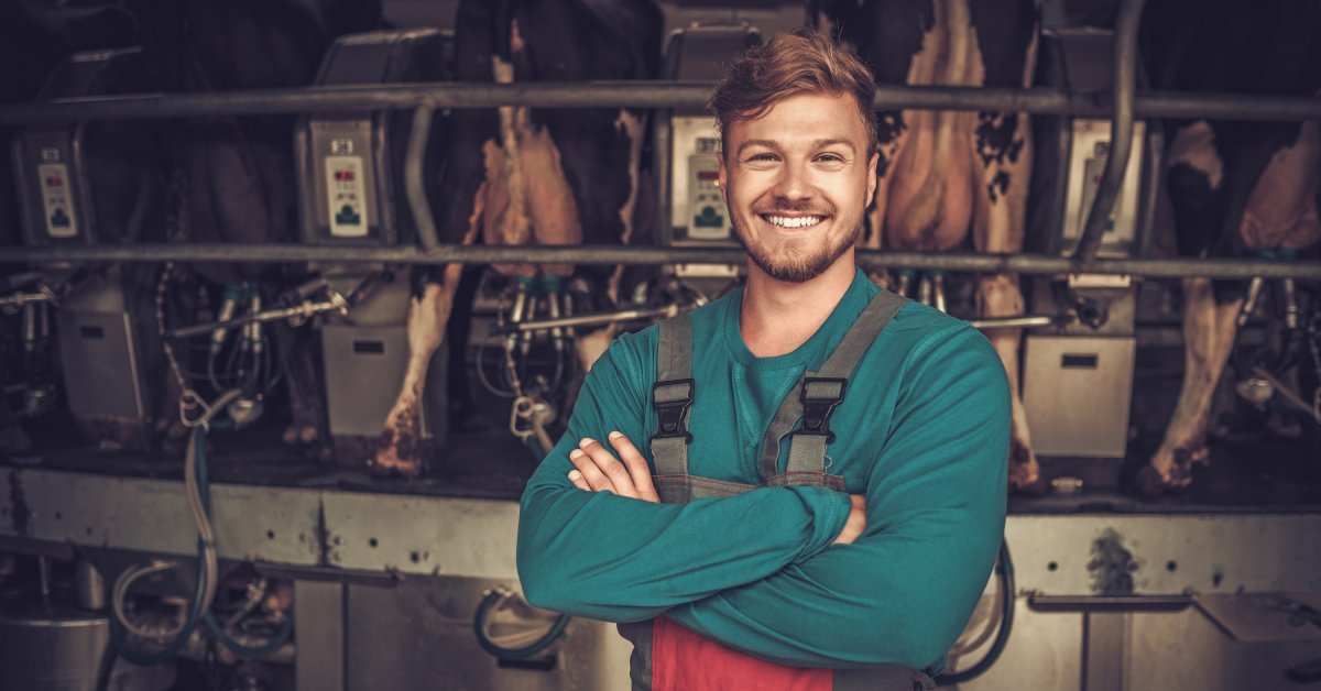 A man in a work apron and long-sleeved shirt smiling. He is standing in front of a row of cows in a miking parlor.