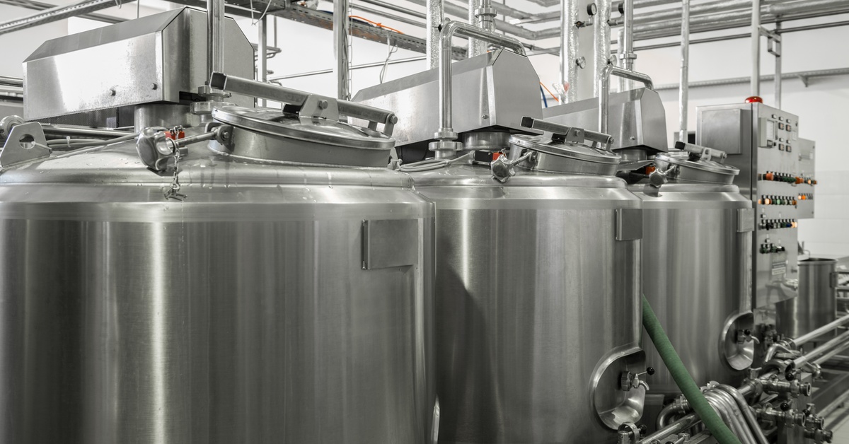 A row of closed stainless-steel bulk milk tanks sits inside a dairy facility, connected by pipes and valves.