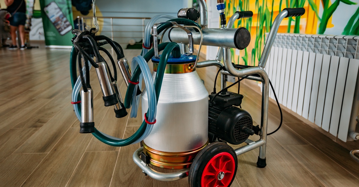 A portable milk bucket unit includes a metal cart frame, hanging hoses, teat cups, and a bucket above red wheels.