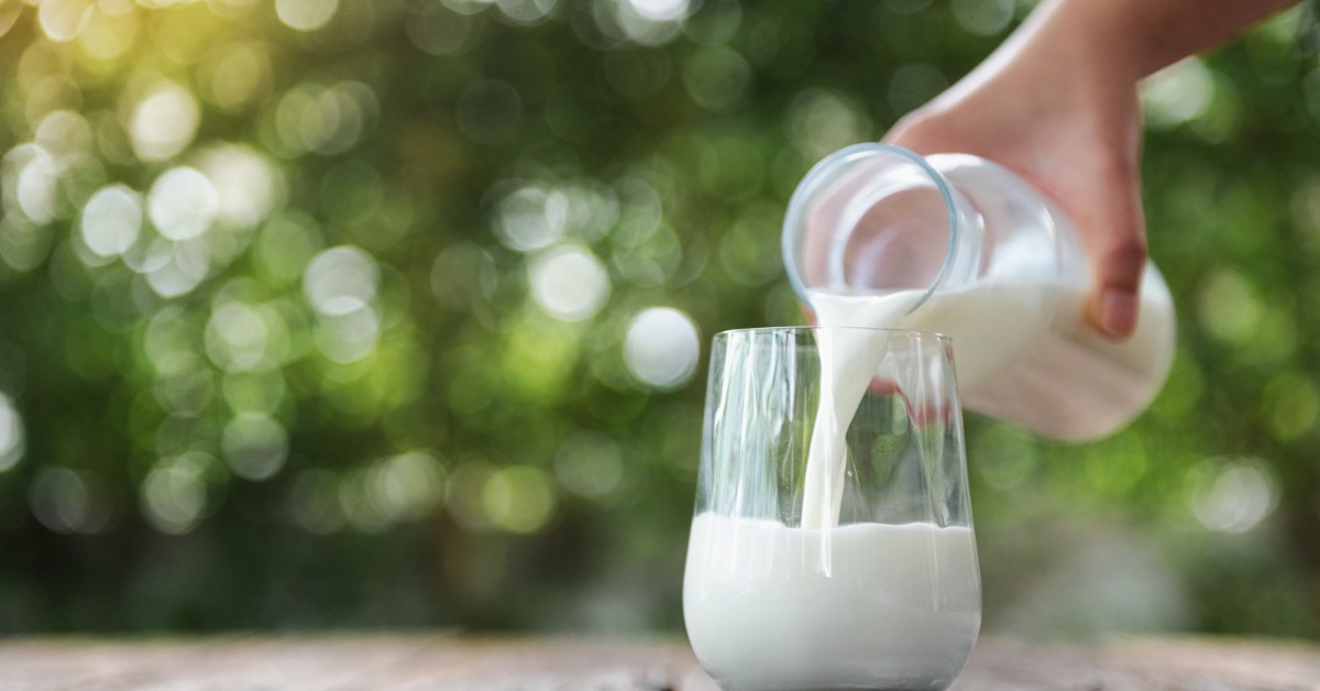 A hand pours milk from a glass bottle into a glass on a wooden table outdoors. Blurred greenery is in the background.