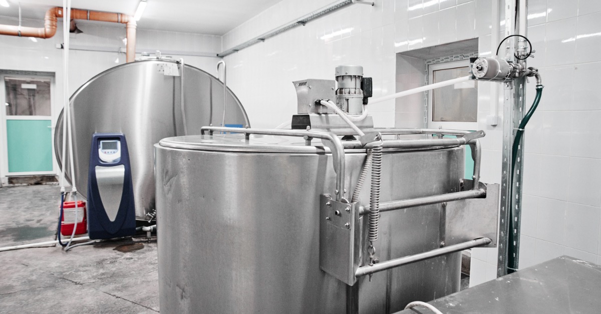 Two large stainless-steel milk tanks, pipes, hoses, motors, and control equipment sit inside a tiled processing room.