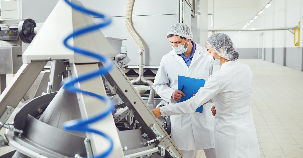 Two workers in white lab coats, masks, and hair nets are inspecting equipment. There is a blue, curled tube in the foreground.