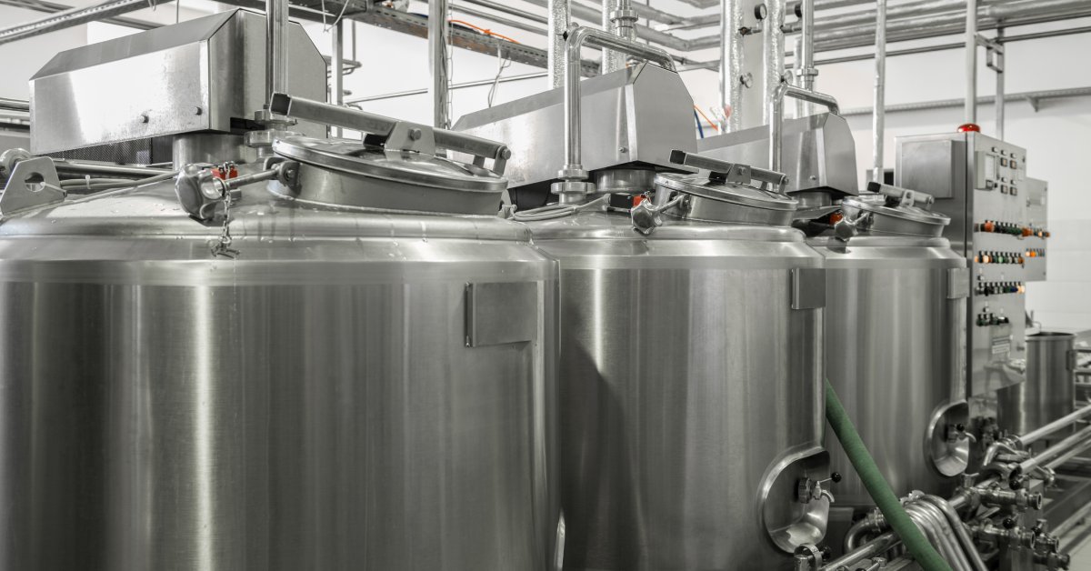 A row of three medium-sized, vertical, metal bulk milk tanks with various pipes, hoses, and gauges connected to them.