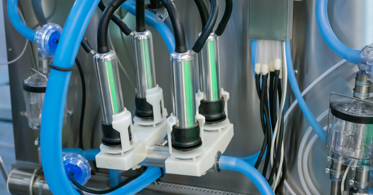 A milking claw with blue tubing and stainless-steel teat cups hung up next to a large metal machine.