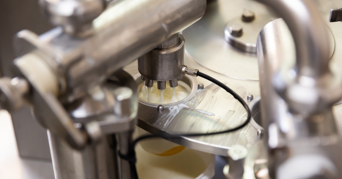 A milk processing machine expelling milk into the opening of a filled tanker as metal parts shine with cleanliness.