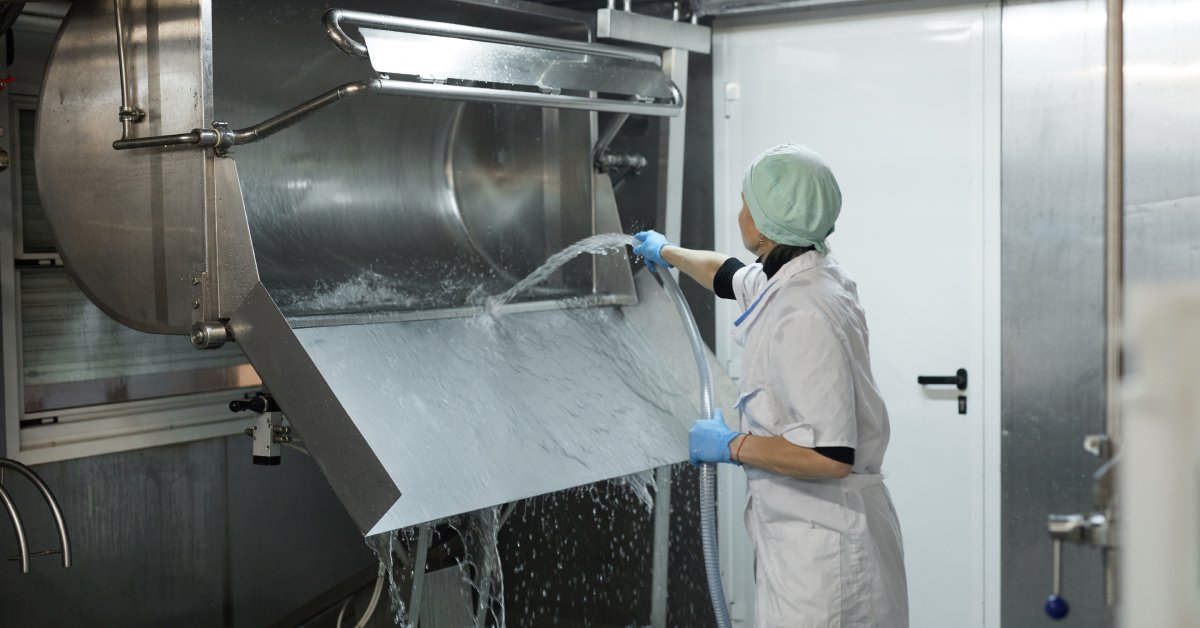 A female worker in a lab coat and hair net is spraying down an open milk cooling tank with a clear hose.
