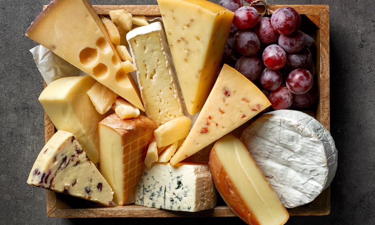 An assortment of cheeses, including Swiss cheese, blue cheese, and brie, arranged in a wooden box with red grapes.
