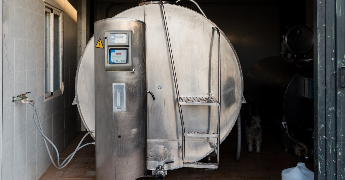 A horizontal metal milk tank stands in a dimly lit indoor space beside a window. The tank's front has a ladder attached.