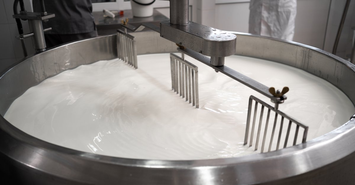 A large, industrial-sized vat of milk is rippling. The machine stirs the milk with two large slotted panels.