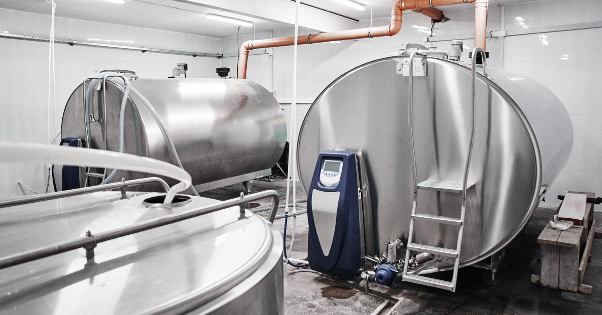 Two large horizontal metal bulk milk tanks, each with a small, three-rung step ladder and a blue control panel.