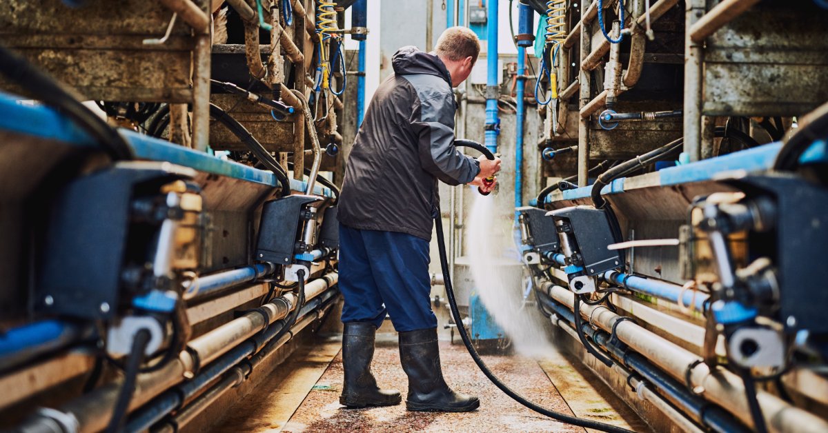 A man wearing a water-resistant jacket and boots using a hose to clean the milking machines between old stables.
